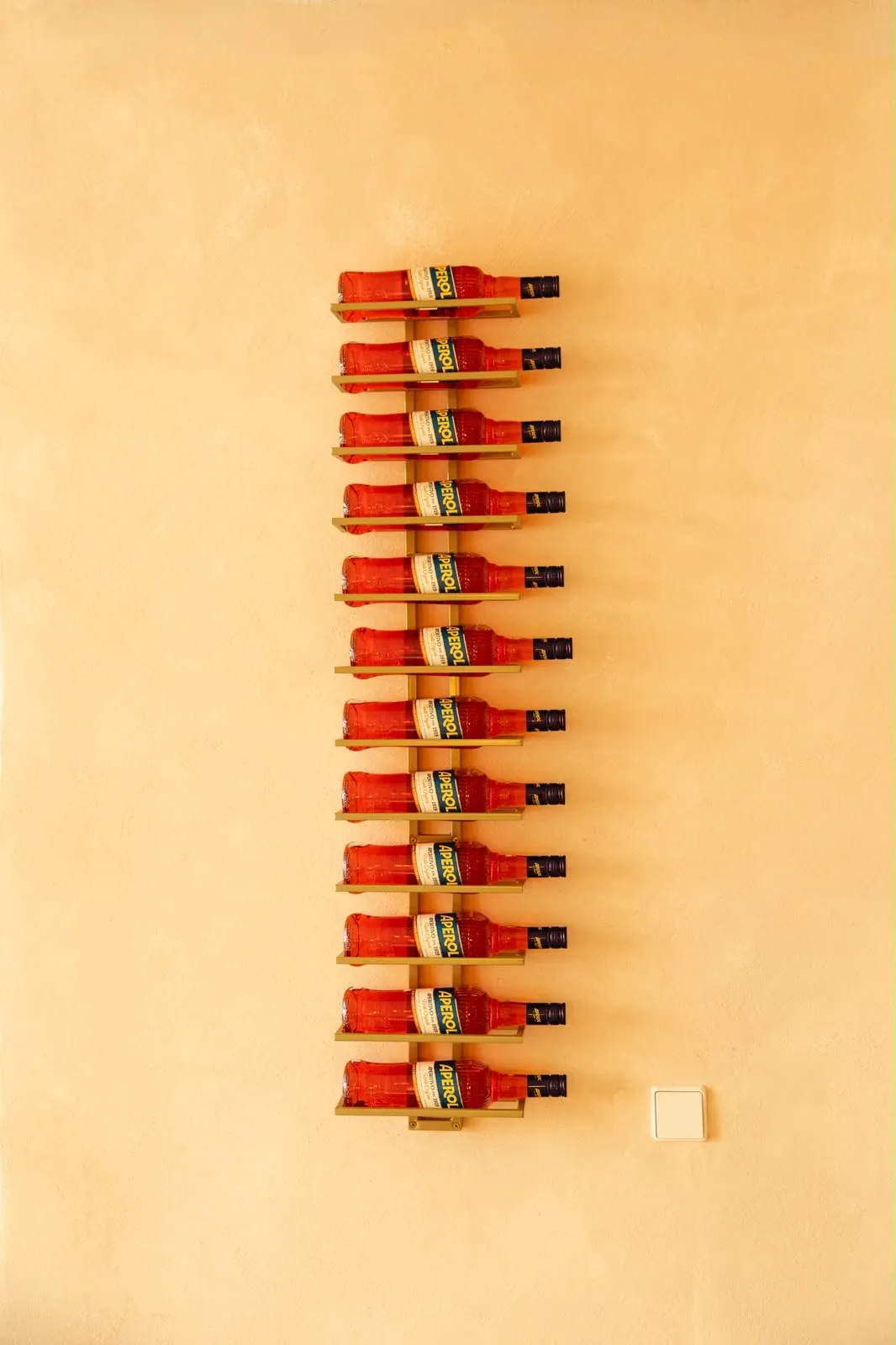 Eine Wandhalterung mit Aperol-Flaschen an der warm getönten Wand.