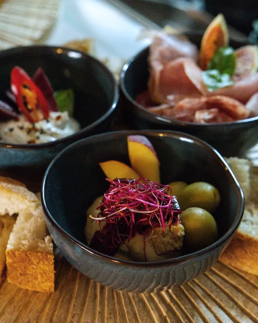 Mezze-Platte auf einem Holzteller mit Schälchen voller Aufschnitt, Frischkäse und Hummus, dazu Brot.