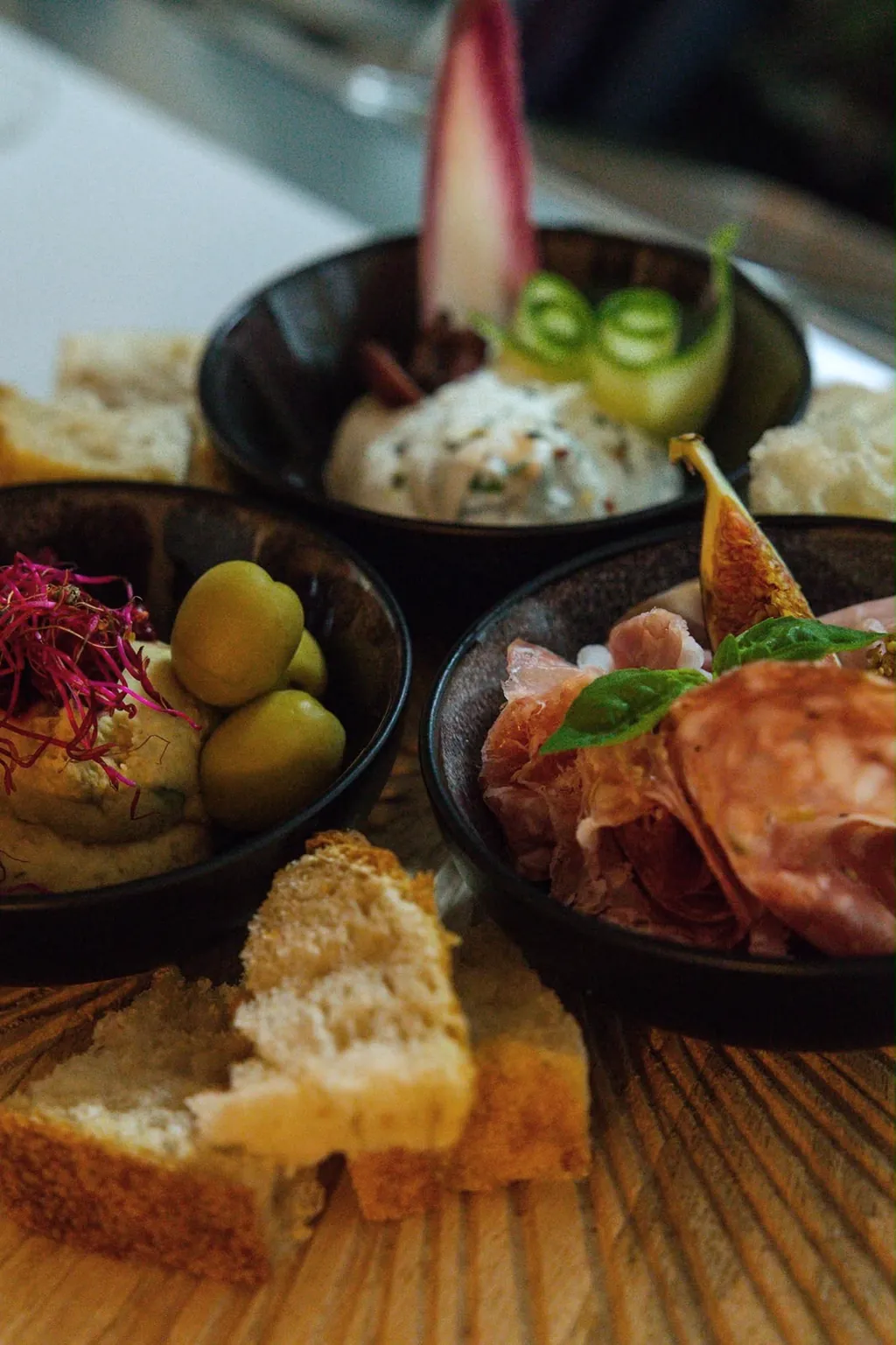 Drei dunkle Schälchen mit Mezze: Frischkäse mit Gurke, Hummus mit Oliven und Aufschnitt mit Feige, davor Brot.