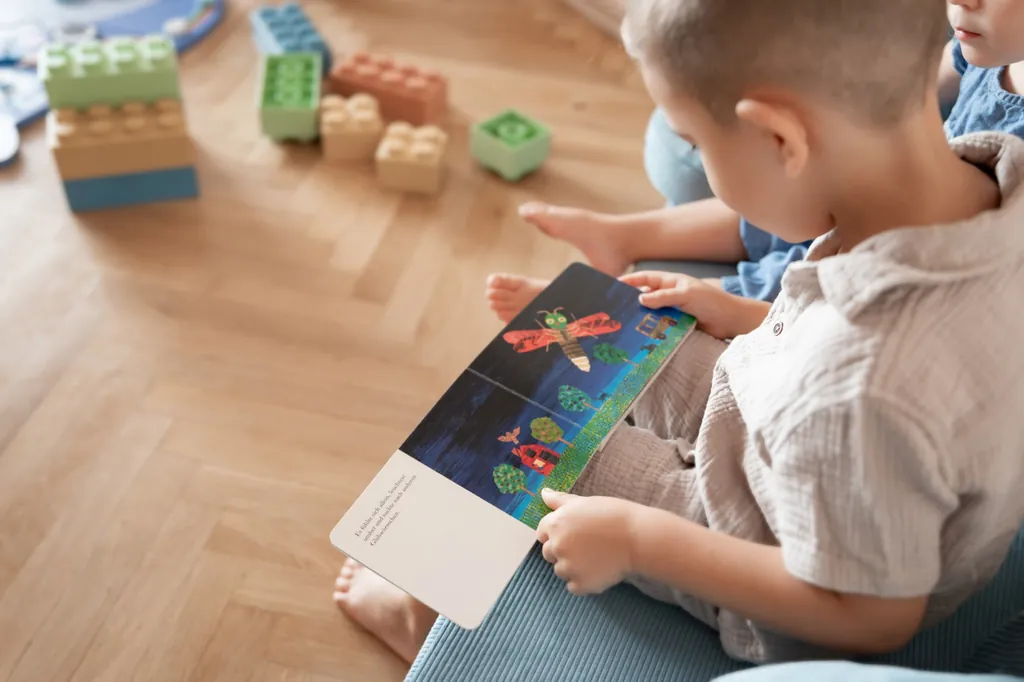 Ein Kind sitzt auf dem Sofa und schaut sich ein Bilderbuch an, auf dem Boden liegen bunte Bausteine.